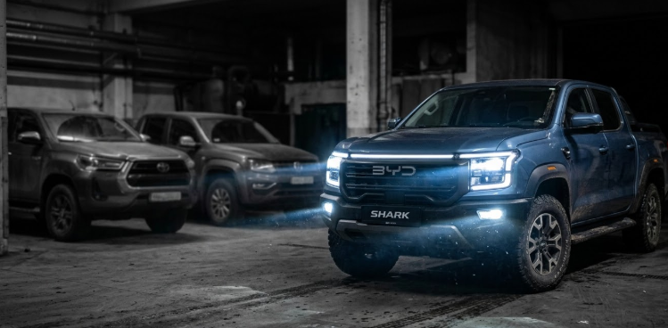 : Una imagen de la BYD Shark emergiendo de la oscuridad con las luces encendidas. Al lado, una Hilux y una Amarok desenfocadas y en blanco y negro".